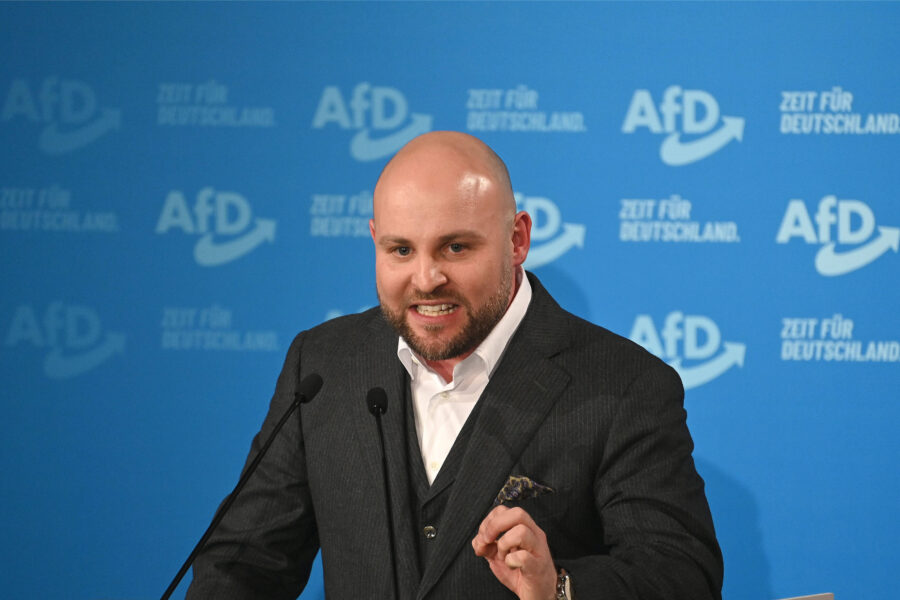 Ein Mann in Anzug spricht an einem Mikrofon vor einer blauen AfD-Wand.