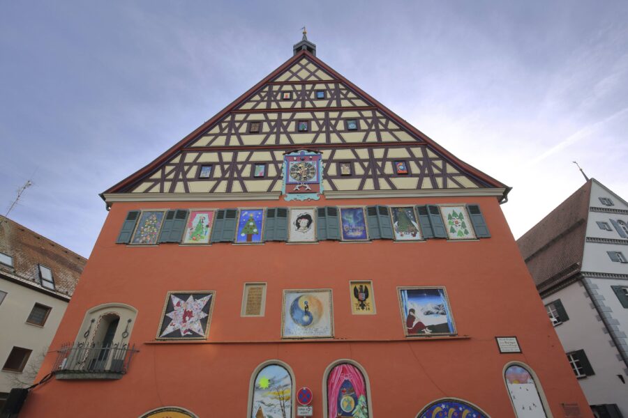Fachwerkhaus mit roter Fassade und bemalten Fenstern, darunter ein Glockenspiel.