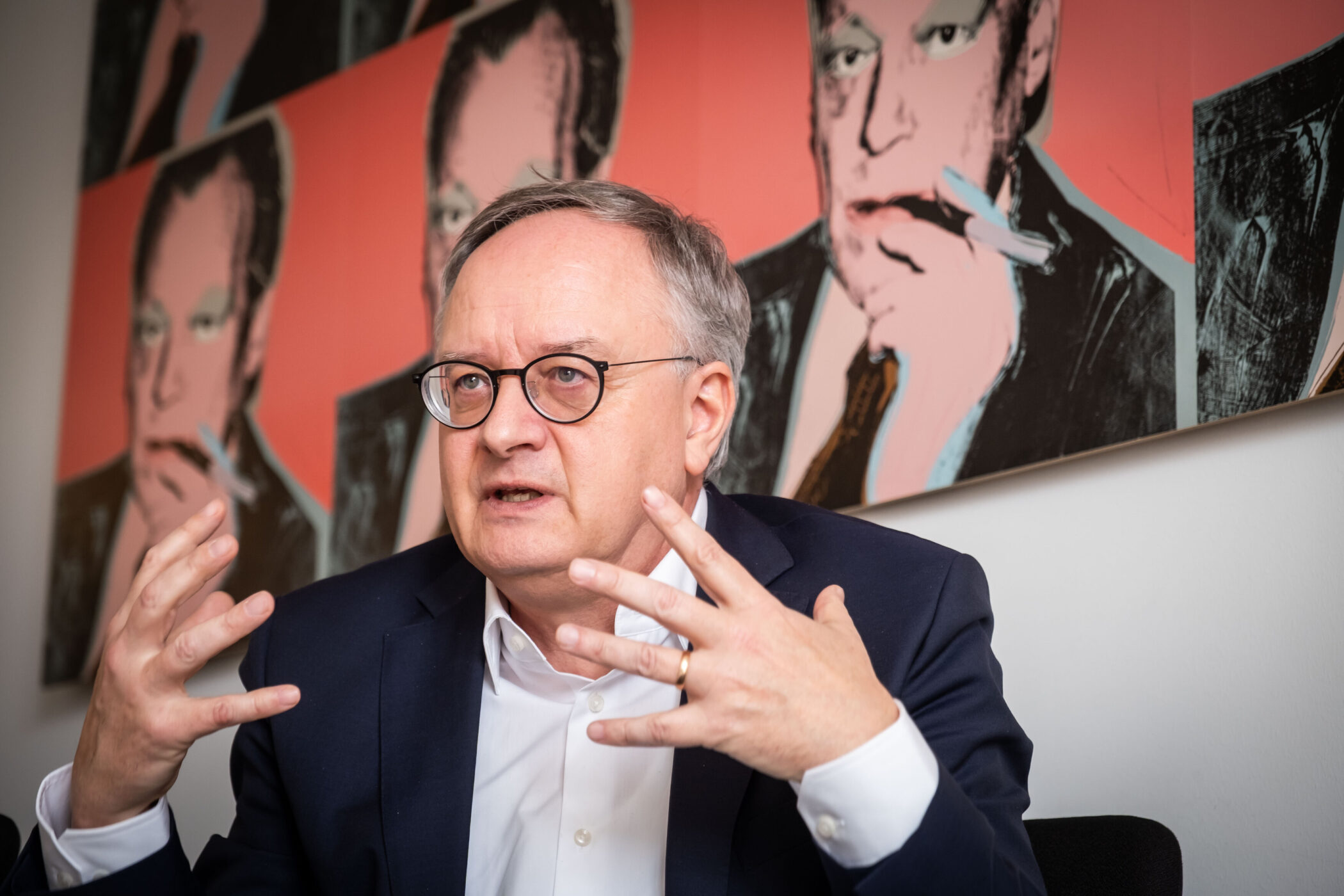 Älterer Mann mit Brille gestikuliert, Hintergrund zeigt ein Porträt mit Zigarette.