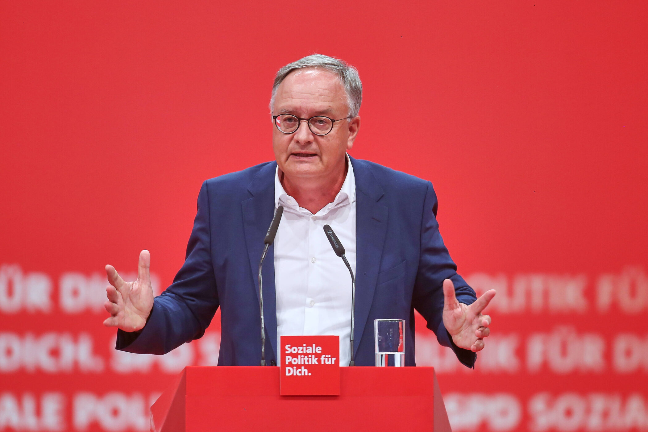 Ein Mann spricht auf roter Bühne mit Mikrofonen und Glas. Text: "Soziale Politik für Dich."