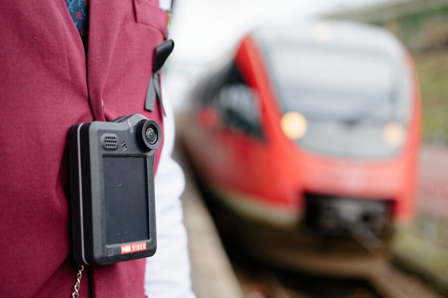 Person mit Bodycam, rote Uniform, roter Zug im Hintergrund.