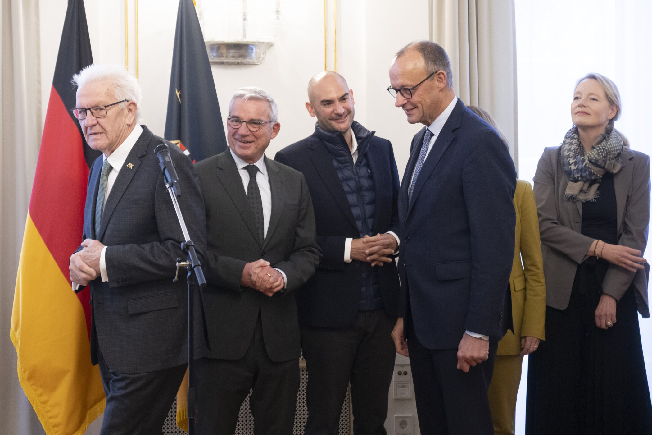 Fünf Personen in Anzügen stehen nebeneinander, zwei Flaggen im Hintergrund.