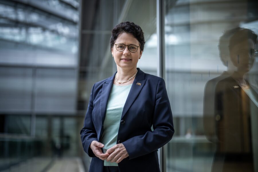 Frau mit kurzer Frisur, Brille, dunklem Blazer vor Glaswand stehend.