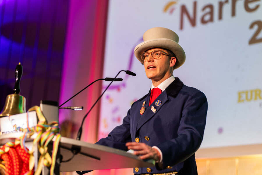 Mann in blauem Anzug und Zylinder spricht an einem Pult mit Mikrofonen.