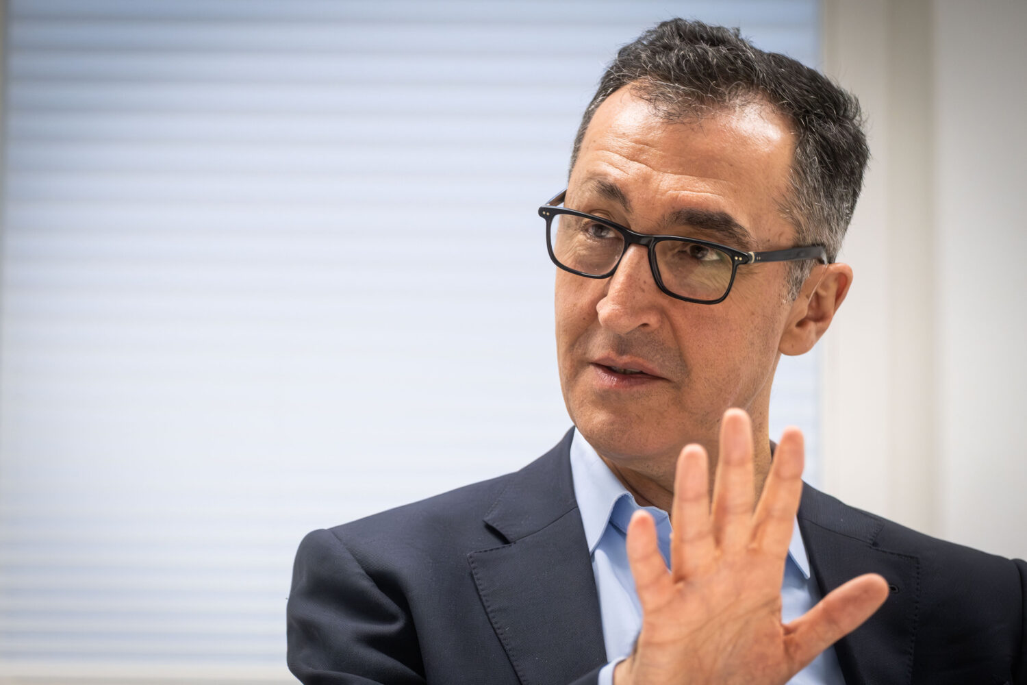 Mann mit Brille und Anzug hebt die Hand, Hintergrund mit Jalousie.