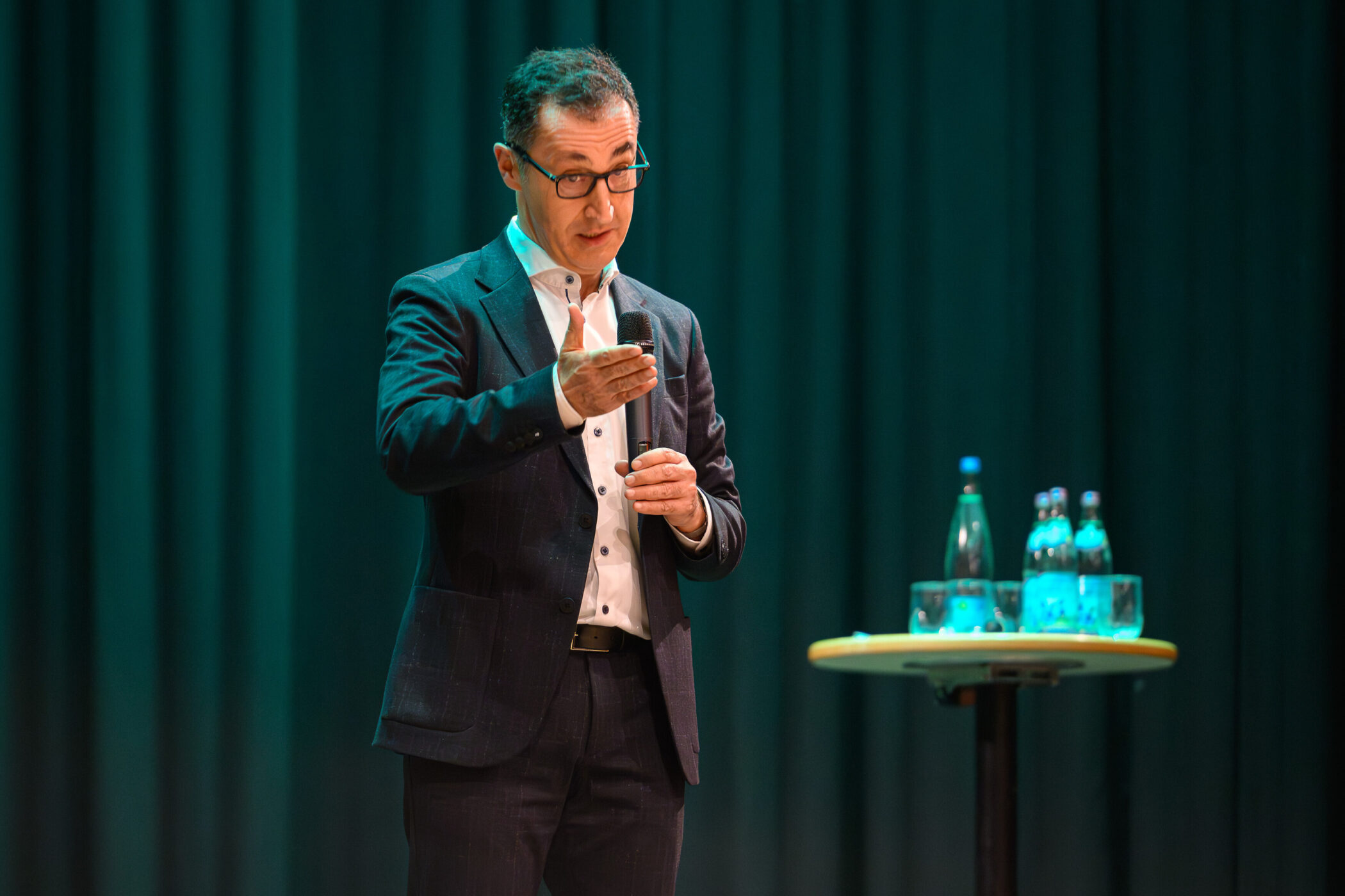 Mann im Anzug spricht mit Mikrofon, Tisch mit Wasserflaschen daneben.