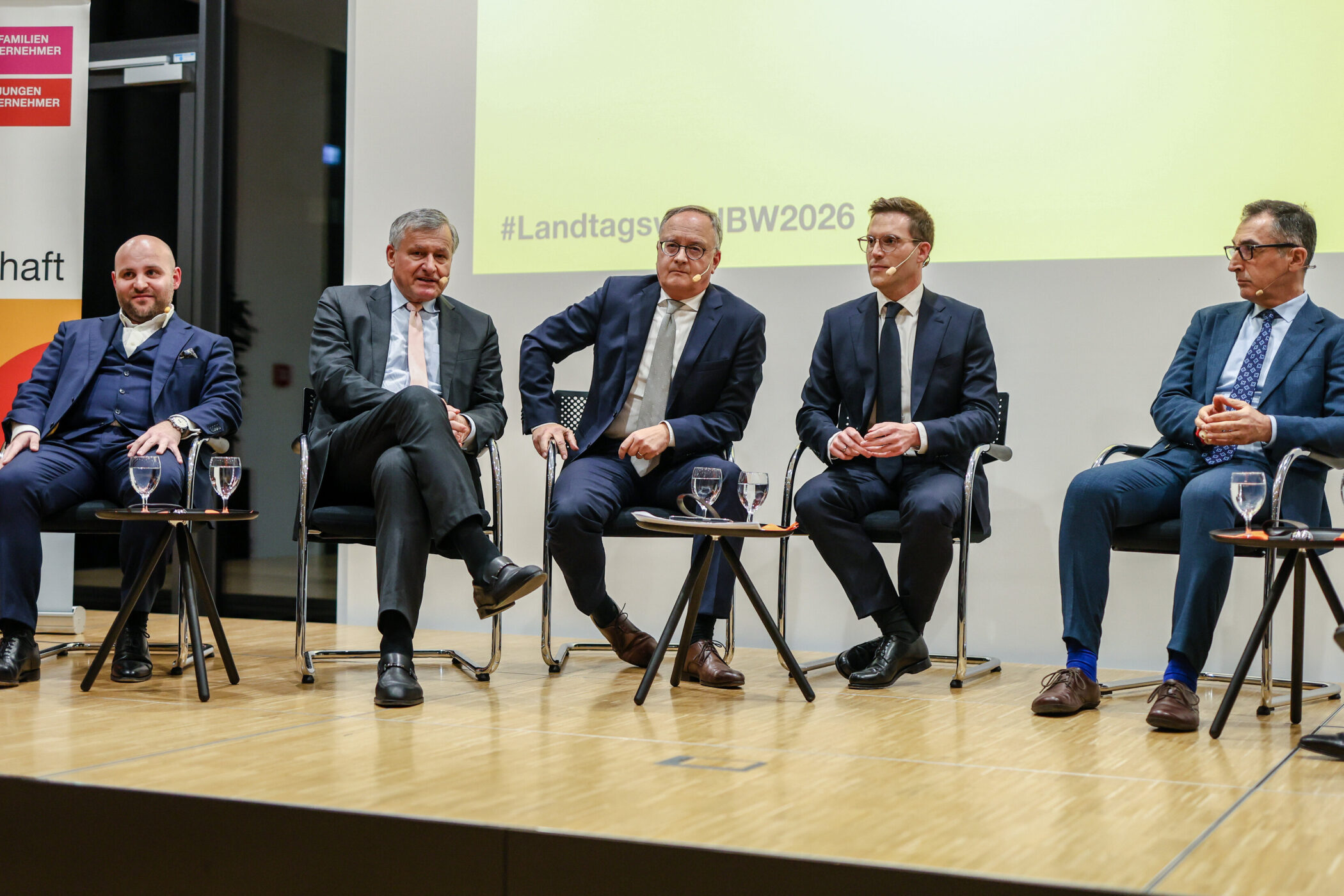 Fünf Männer im Anzug sitzen auf einer Bühne neben Tischen mit Wasser.
