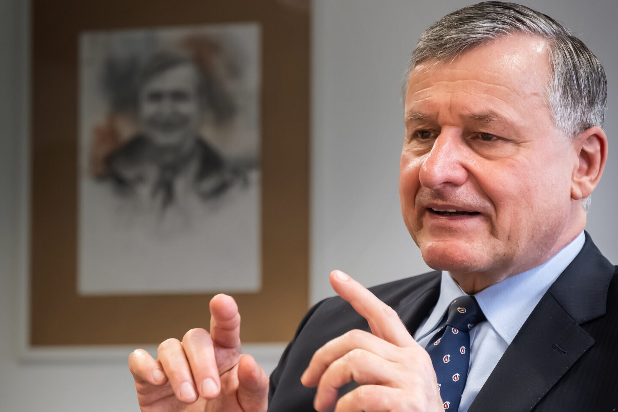 Älterer Mann im Anzug gestikuliert, im Hintergrund ein unscharfes Porträt.