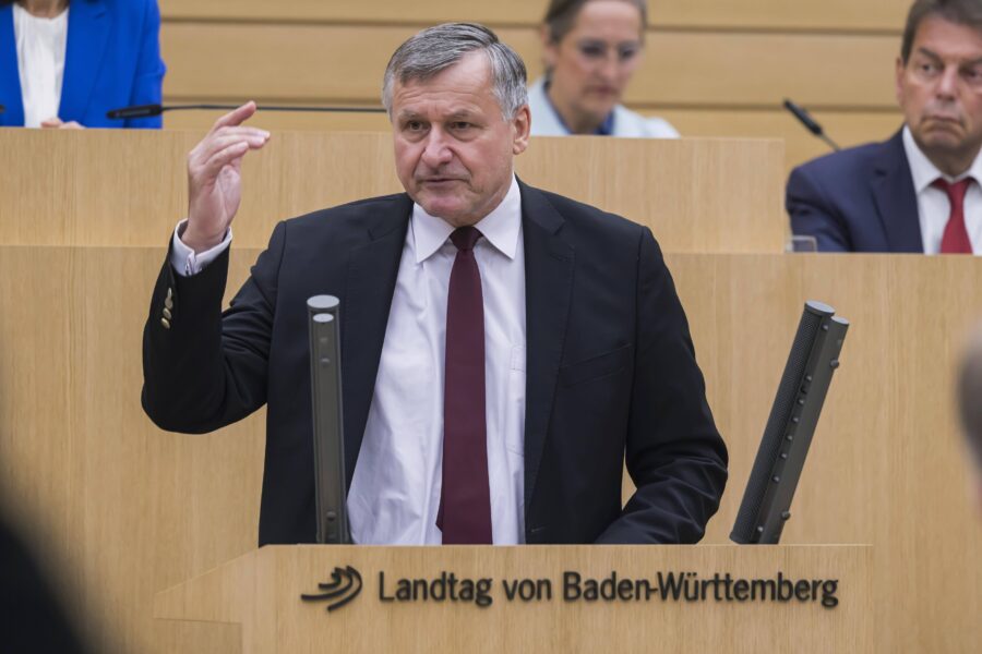 Mann im Anzug spricht im Landtag von Baden-Württemberg, zwei Personen im Hintergrund.
