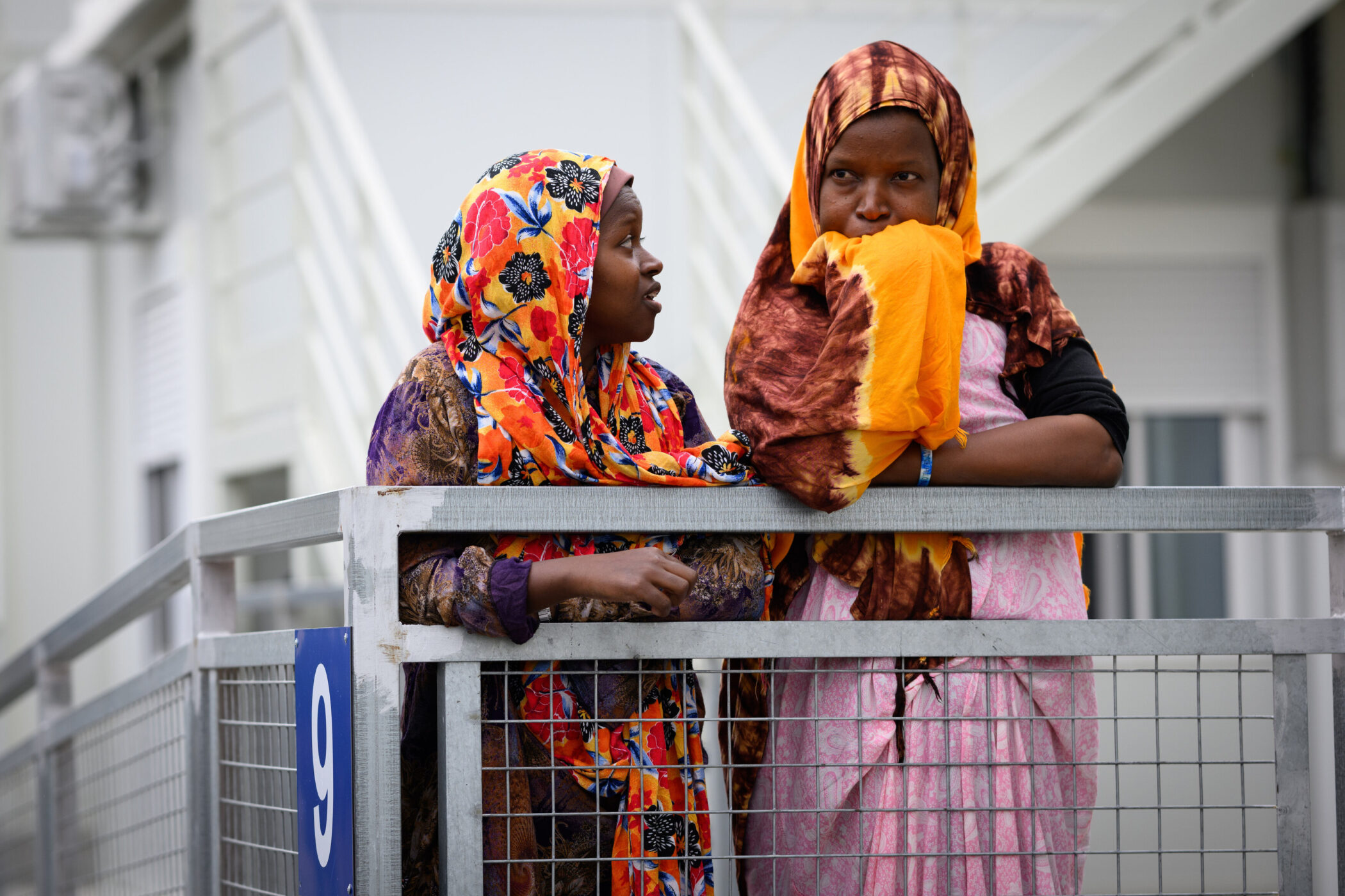 Zwei Frauen in bunten Kopftüchern stehen an einem Geländer, Nummer 9 sichtbar.