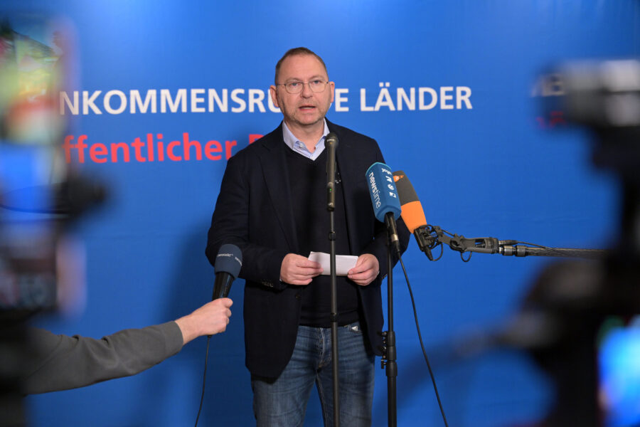 Mann hält Ansprache hinter Mikrofonen vor blauem Hintergrund mit Schrift.