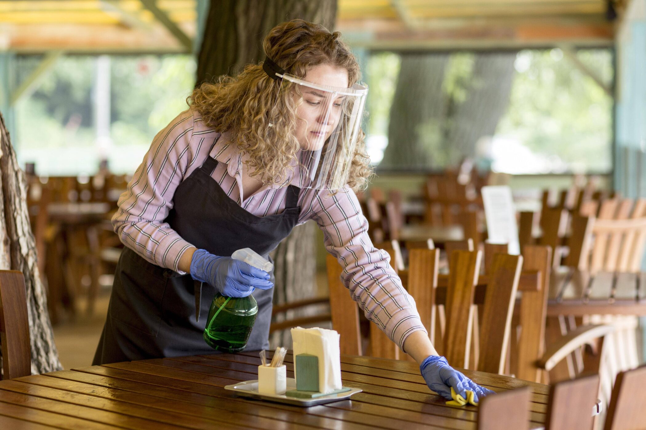 Person mit Gesichtsschutz reinigt einen Holztisch in einem Restaurant.