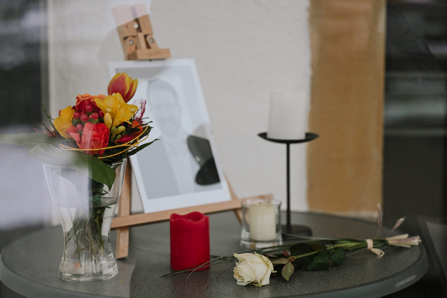 Gerahmtes Foto auf Staffelei, Blumenstrauß, Kerzen und weiße Rose auf Tisch.