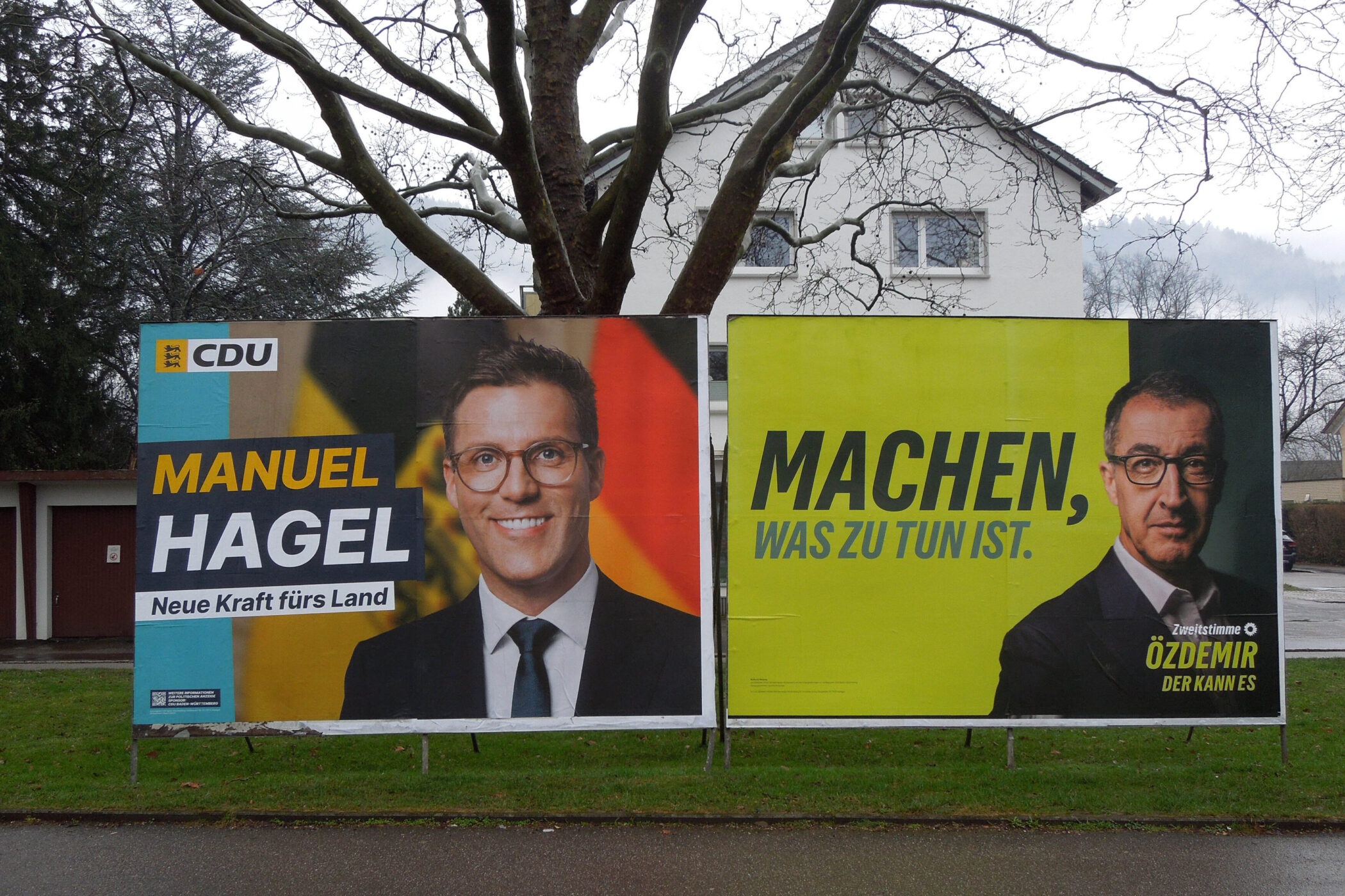 Zwei Wahlplakate nebeneinander auf einer regennassen Straße.