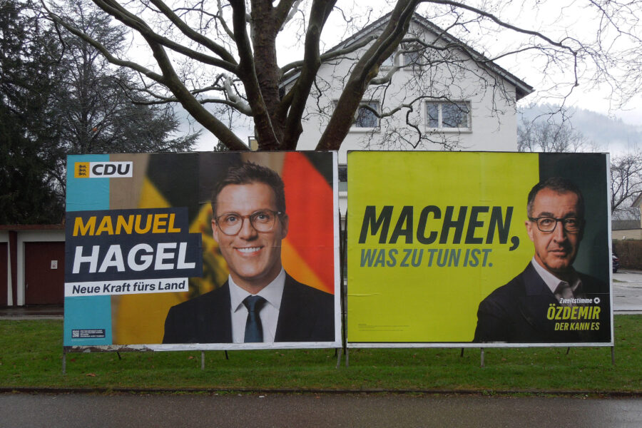 Zwei Wahlplakate nebeneinander auf einer regennassen Straße.