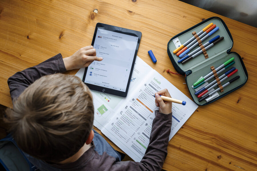 Kind mit Tablet und Heft, ein Federmäppchen mit Stiften auf dem Tisch.