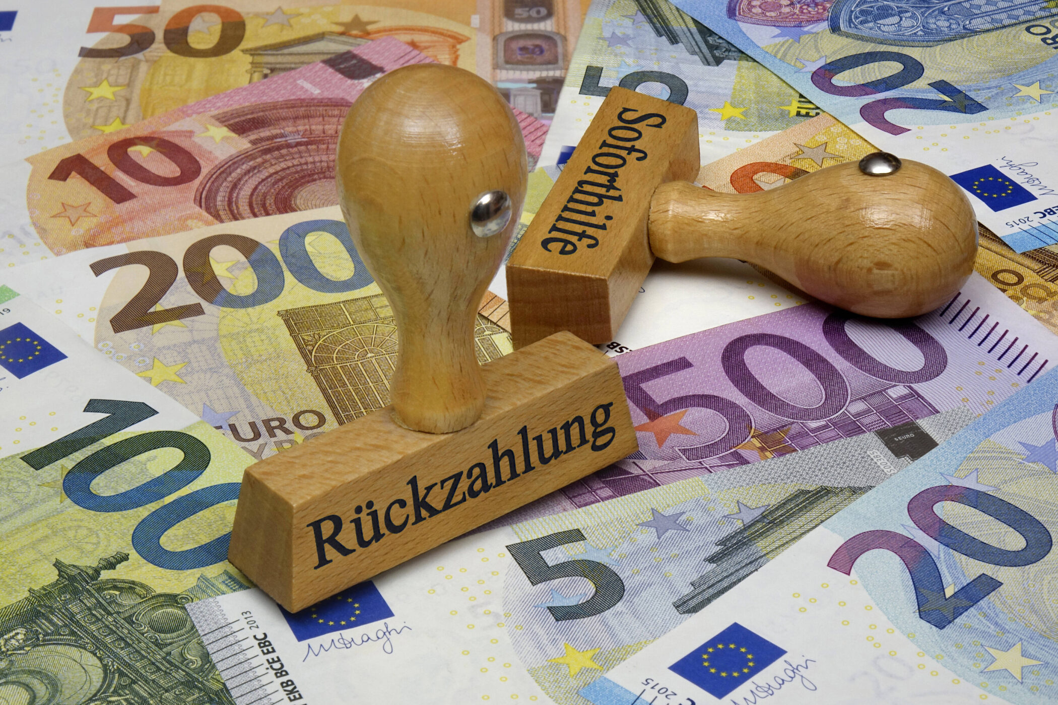 Eurobanknoten und zwei Holzstempel mit "Rückzahlung" und "Soforthilfe".