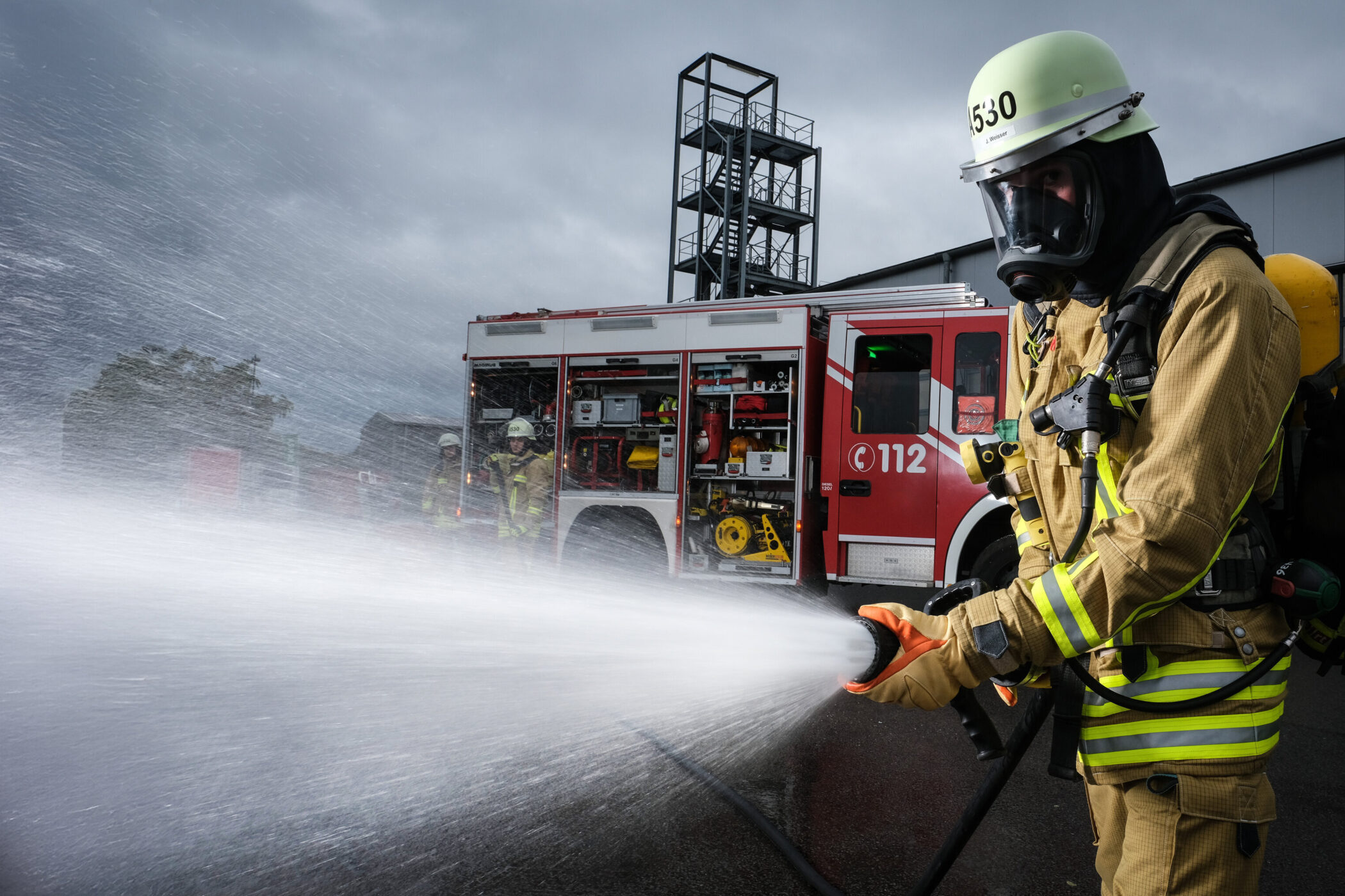 Feuerwehrmann in Schutzkleidung spritzt Wasser, Feuerwehrauto im Hintergrund.
