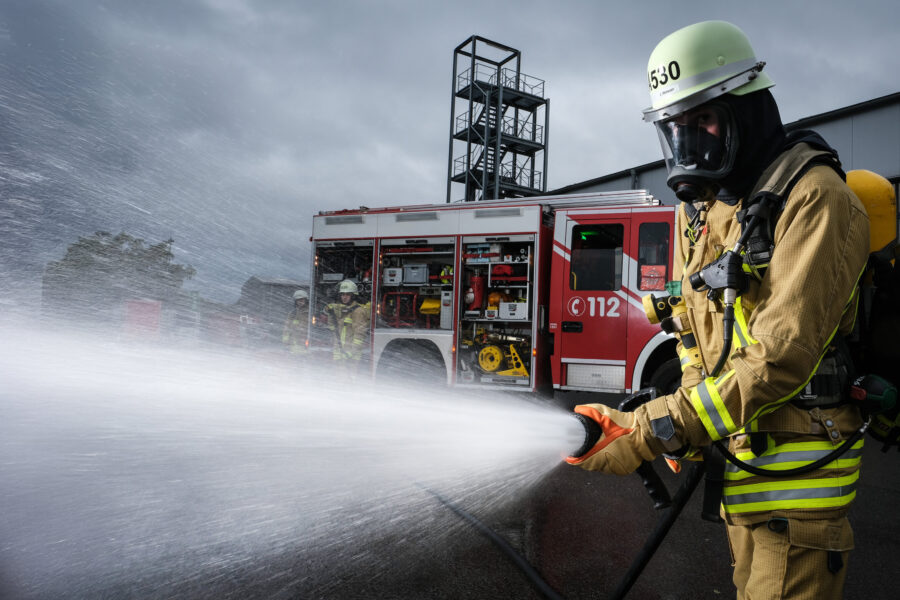 Feuerwehrmann in Schutzkleidung spritzt Wasser, Feuerwehrauto im Hintergrund.
