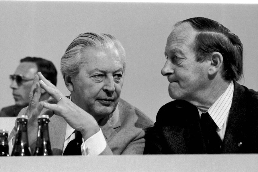 Drei Männer in Anzügen, zwei im Gespräch, Flaschen im Vordergrund. Schwarz-Weiß-Foto.