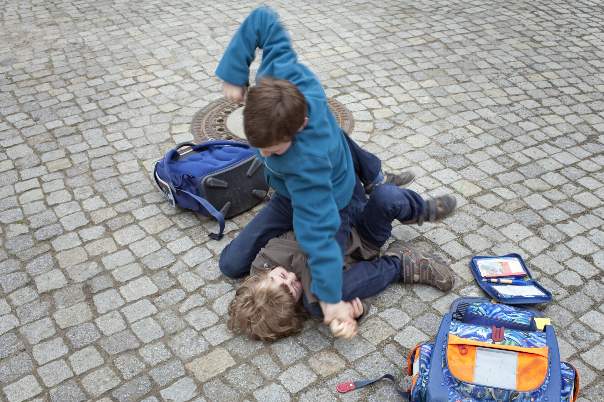 Zwei Jungen raufen auf einem gepflasterten Boden, Rucksäcke liegen daneben.