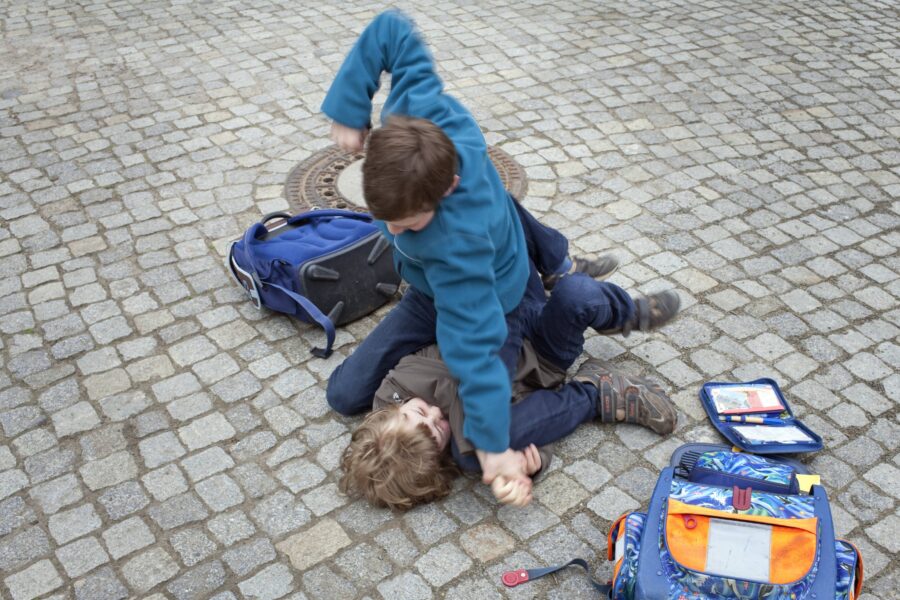 Zwei Jungen raufen auf einem gepflasterten Boden, Rucksäcke liegen daneben.