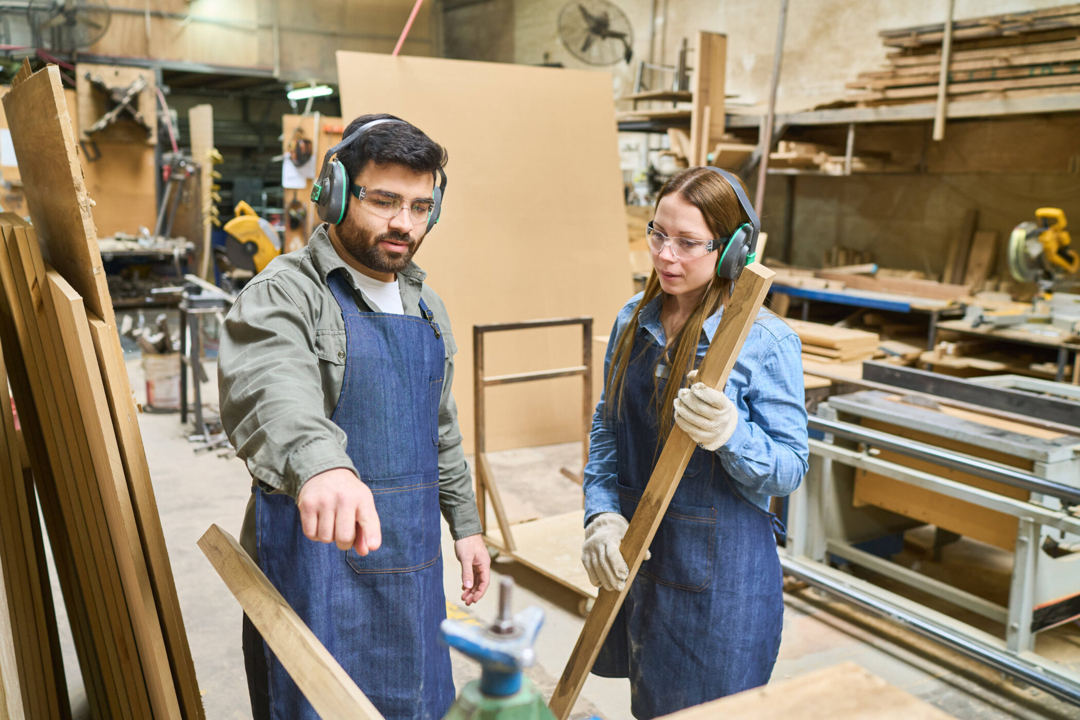 Zwei Personen mit Gehörschutz arbeiten in einer Werkstatt, eine hält ein Holzbrett.