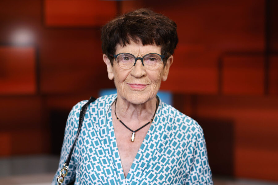 Ältere Frau mit Brille und gemustertem Oberteil vor unscharfem Hintergrund.