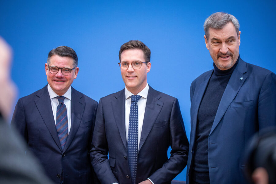 Drei Männer in Anzügen vor blauem Hintergrund.