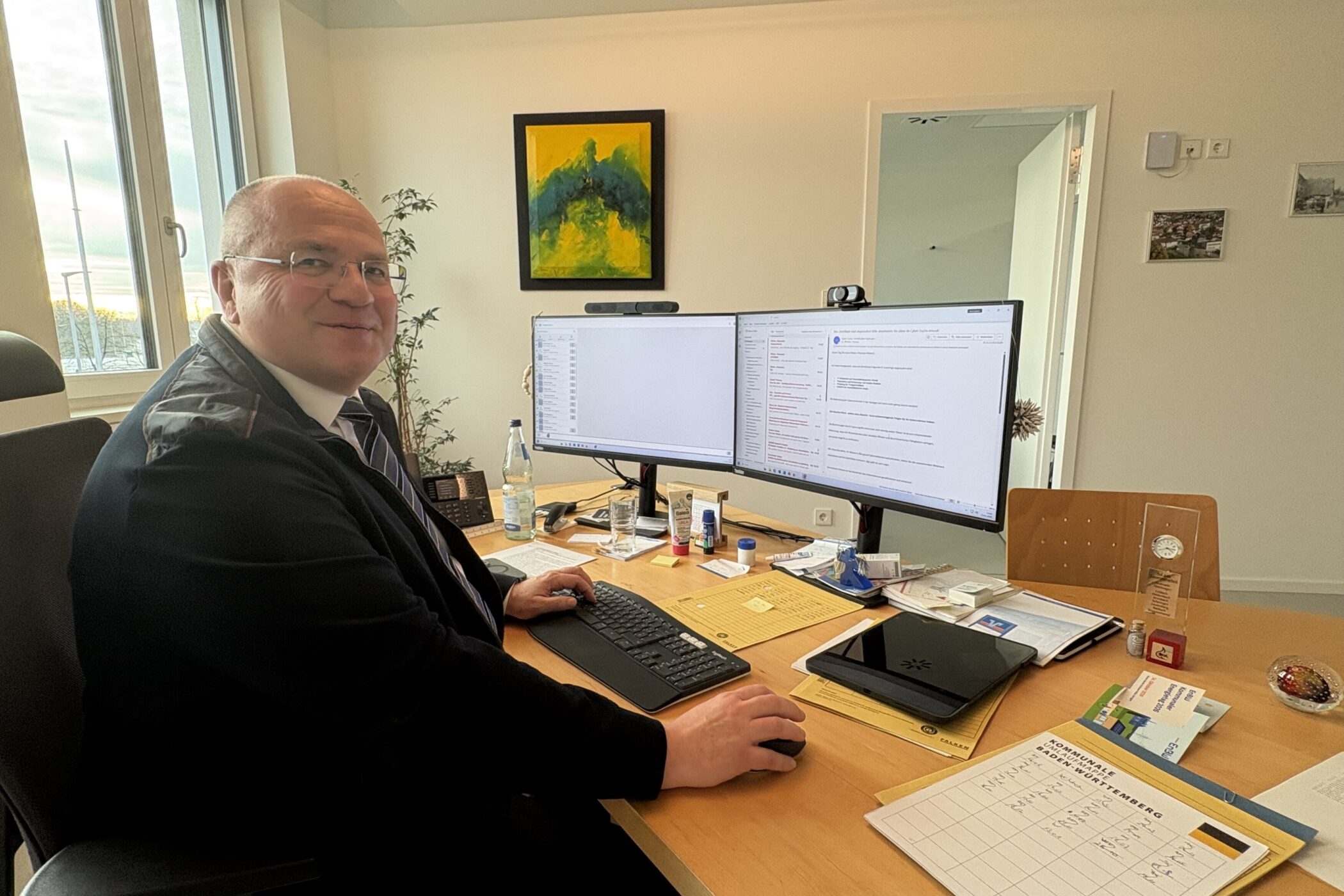 Mann am Schreibtisch mit zwei Monitoren, Tastatur und Büromaterialien.