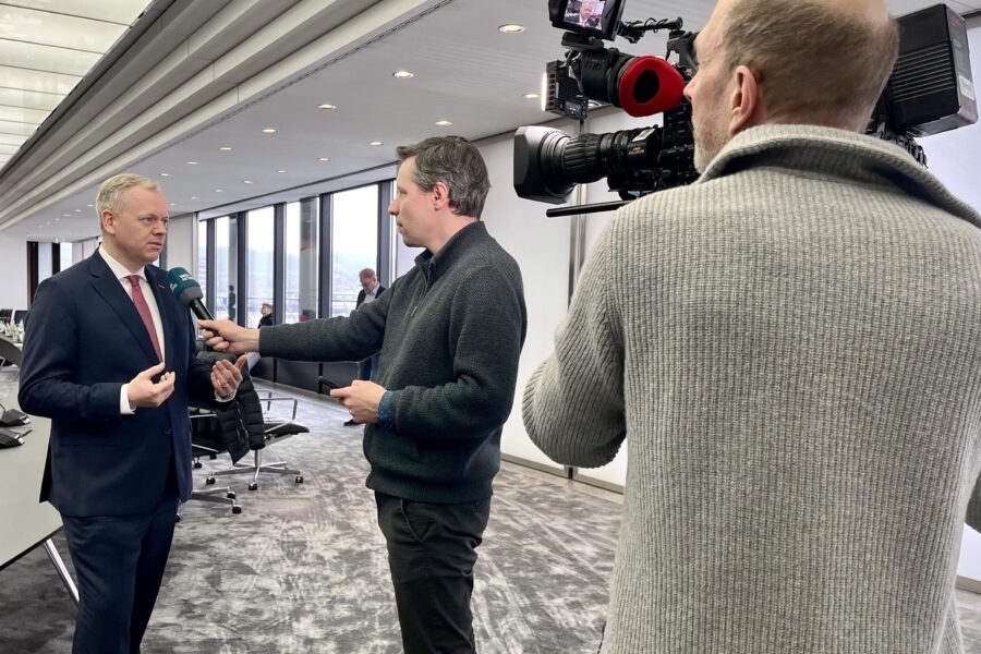 Drei Männer in einem Büro, einer wird interviewt, ein Kameramann filmt.
