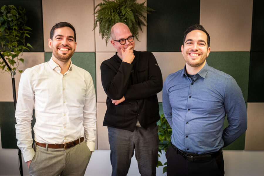 Drei Männer stehen lächelnd vor einer Wand mit Pflanzen.