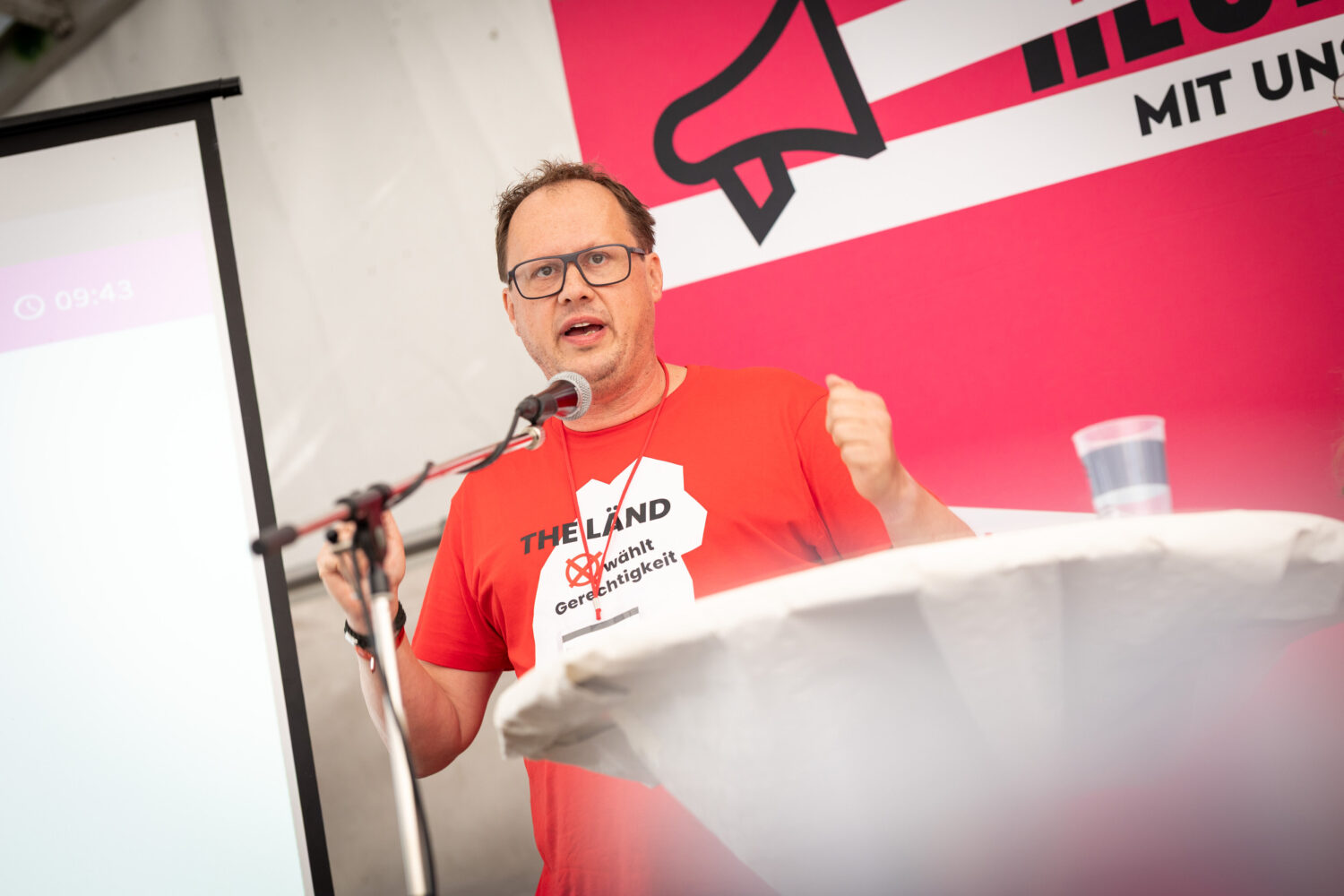 Mann in rotem T-Shirt spricht am Mikrofon, daneben Leinwand und roter Hintergrund.