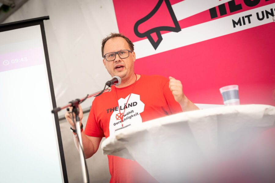 Mann in rotem T-Shirt spricht am Mikrofon, daneben Leinwand und roter Hintergrund.