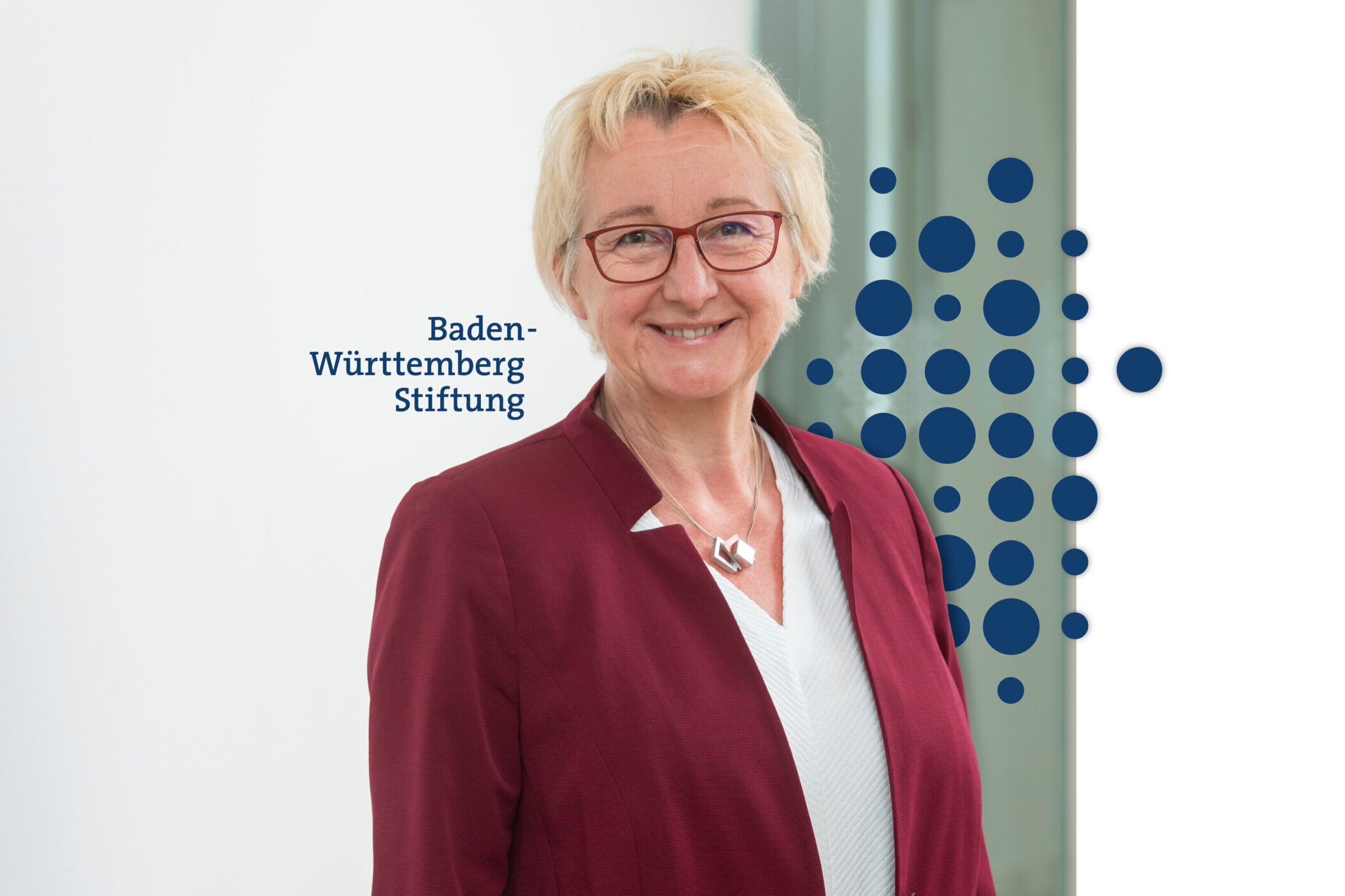 Ältere Frau mit Brille in roter Jacke lächelt. Text: „Baden-Württemberg Stiftung”.