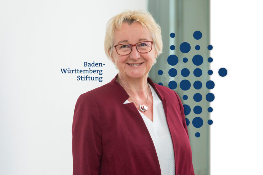 Ältere Frau mit Brille in roter Jacke lächelt. Text: „Baden-Württemberg Stiftung”.