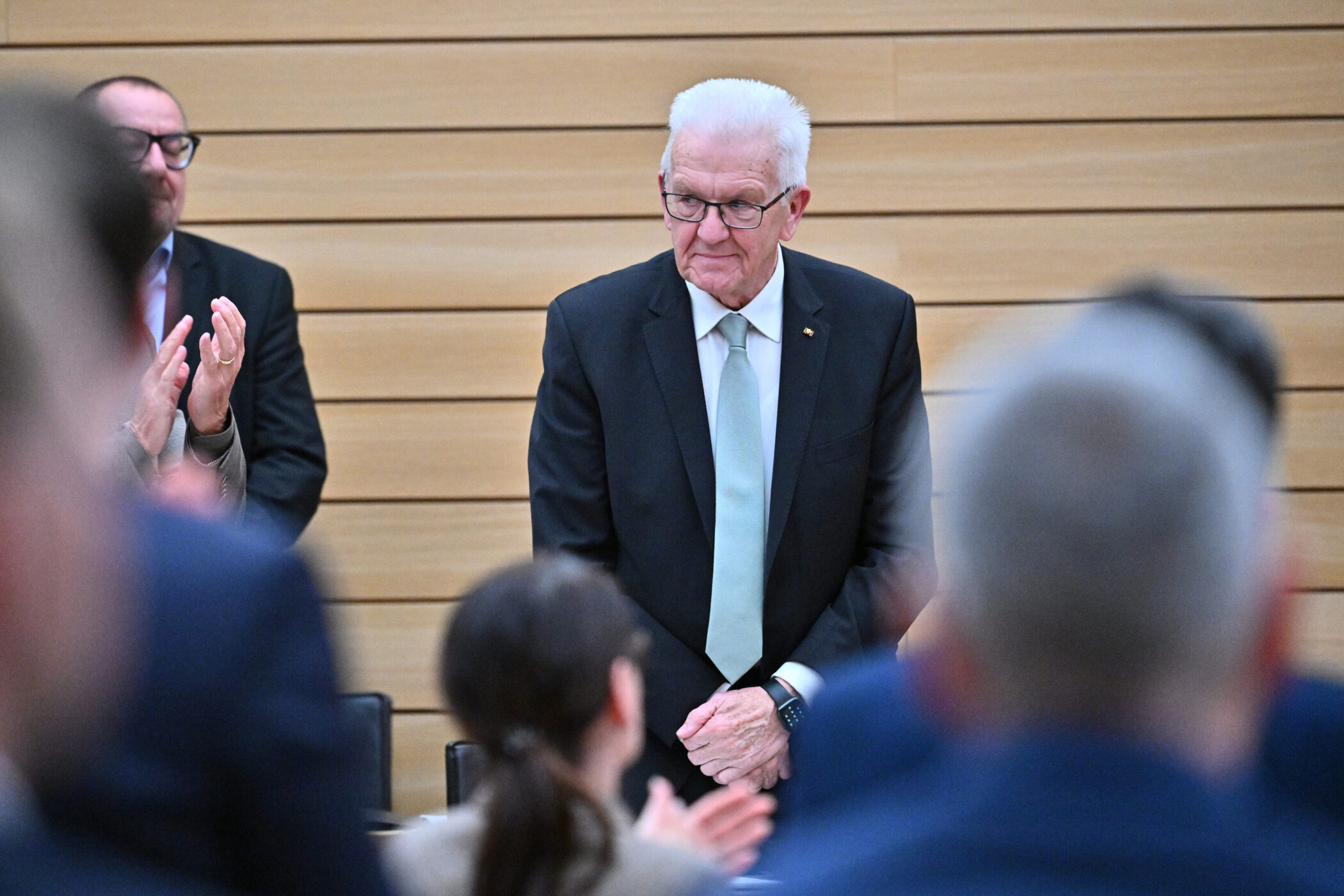 Älterer Mann im Anzug steht, umgeben von klatschenden Menschen.