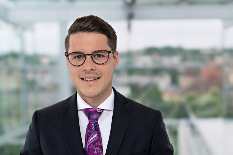 Mann mit Brille im Anzug, bunter Krawatte, unscharfer Hintergrund.