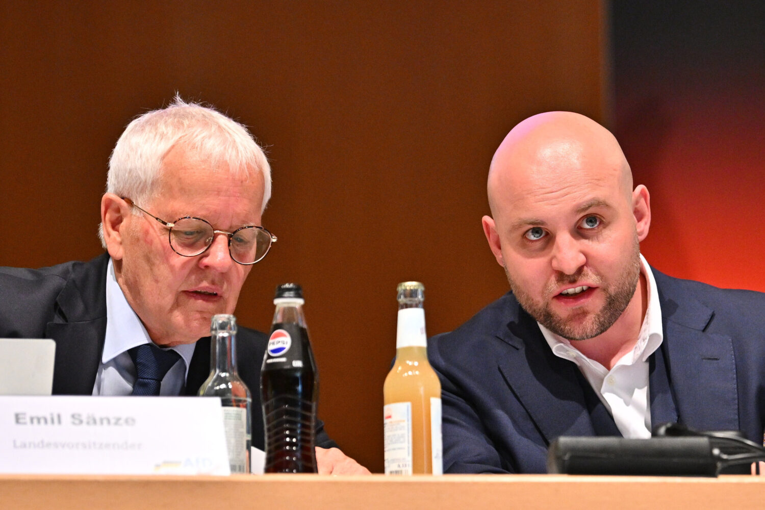 Zwei Männer sitzen an einem Tisch mit Getränkeflaschen. Ein Namensschild ist sichtbar.