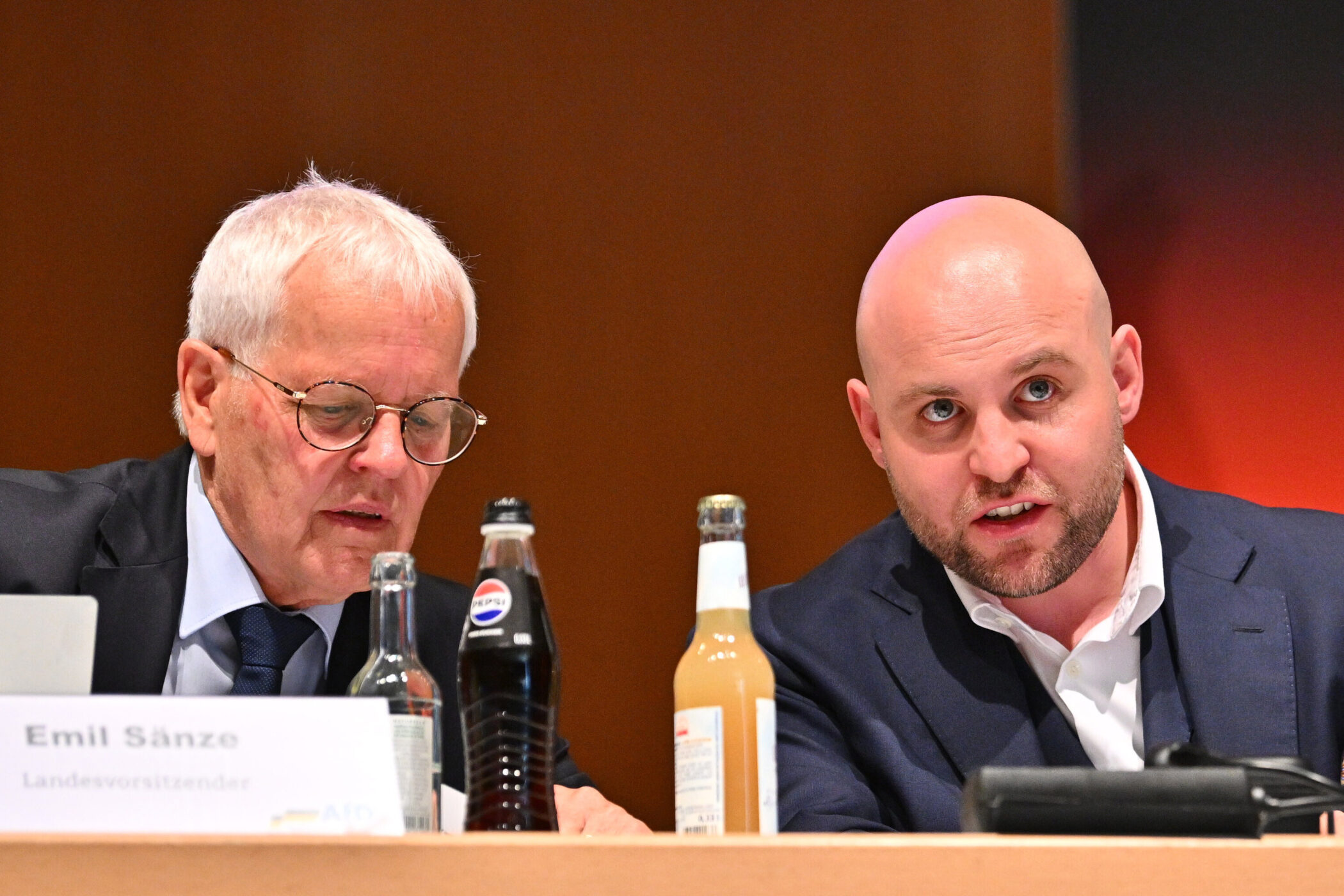 Zwei Männer sitzen an einem Tisch mit Getränkeflaschen. Ein Namensschild ist sichtbar.