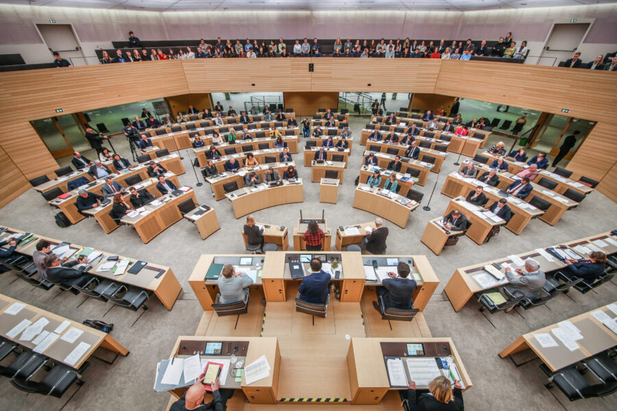 Versammlung im großen Hörsaal mit Teilnehmern an Holztischen, Publikum im Hintergrund.