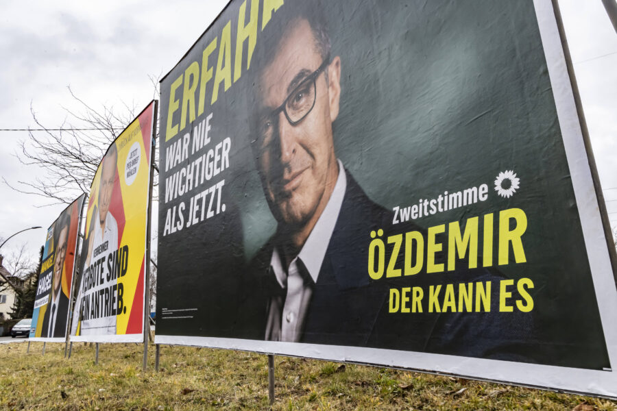 Mehrere Wahlplakate an einer Straße, teils mit Text und Porträts.