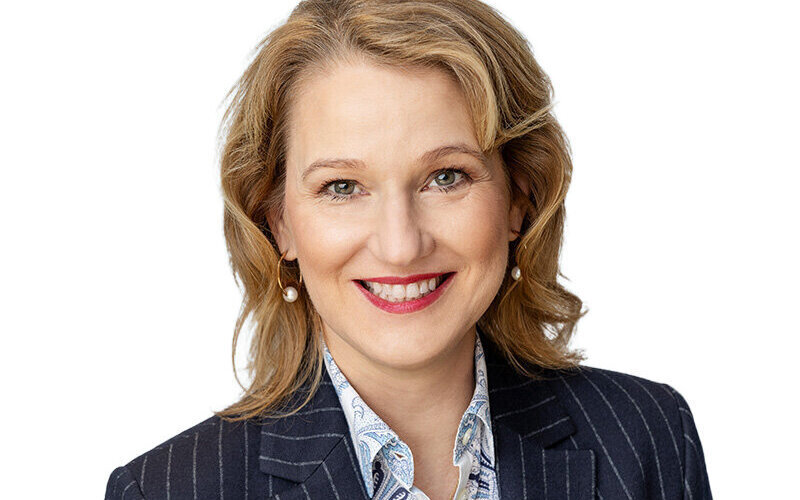 Person mit blonden Haaren und gestreiftem Anzug lächelt vor weißem Hintergrund.