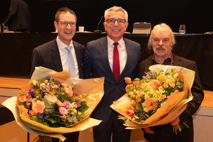 Drei Männer in Anzügen halten Blumensträuße und lächeln in die Kamera.