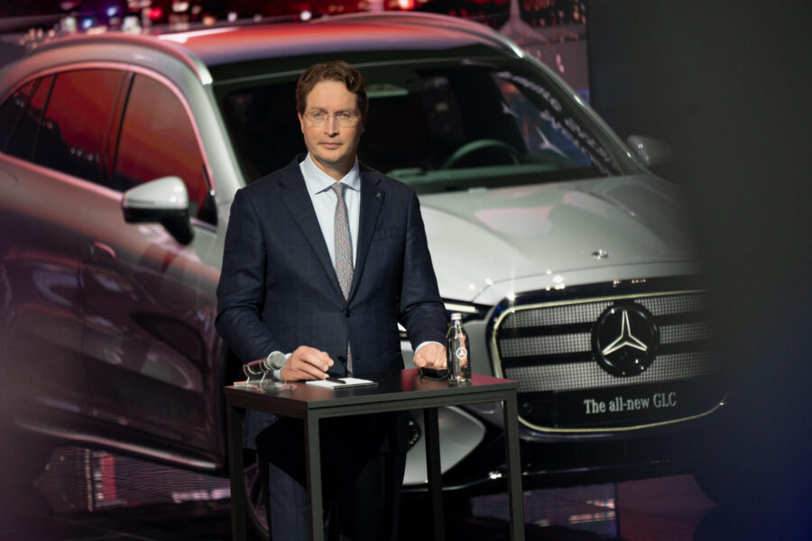 Mann im Anzug steht vor einem silbernen Mercedes GLC mit Mikrofon und Wasserflasche.