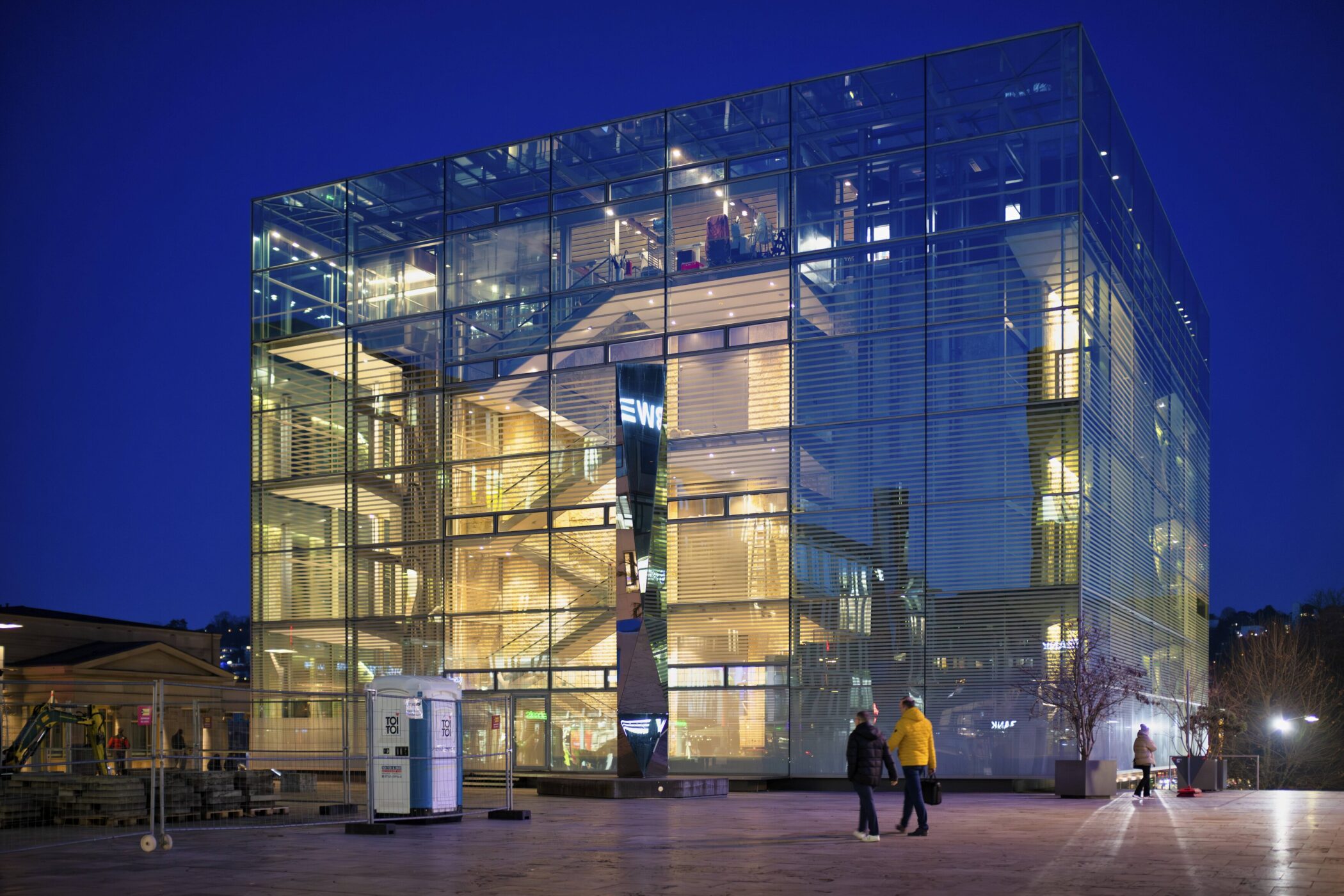 Glasgebäude bei Nacht, zwei Personen gehen vorbei, zwei mobile Toiletten im Vordergrund.