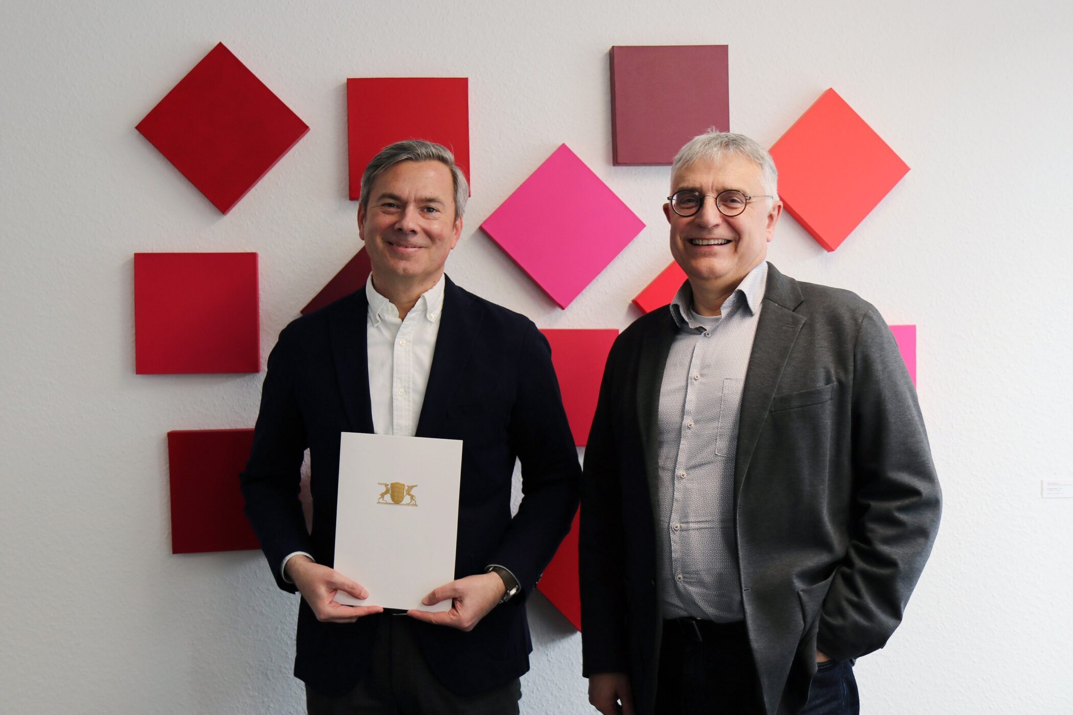 Zwei Männer vor einer Wand mit roten Quadraten, einer hält ein Dokument.