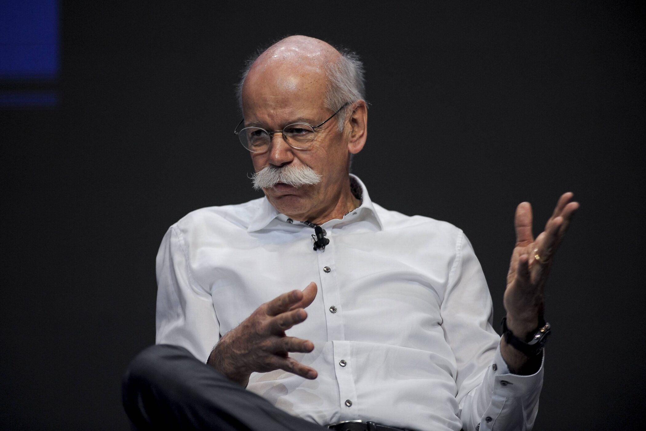 Älterer Mann mit Brille und Schnurrbart in weißem Hemd gestikuliert.