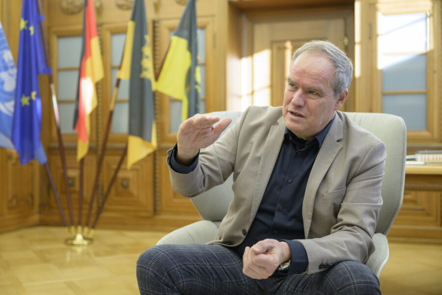 Ein Mann im Anzug gestikuliert sitzend, im Hintergrund mehrere Flaggen.