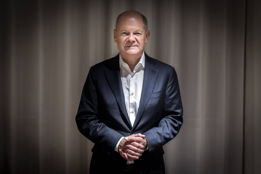 Ein Mann in Anzug steht vor einem Vorhang, die Hände gefaltet.